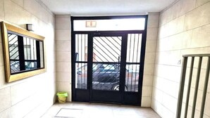 A hallway with a tiled floor, a black door with a window, and a yellow trash bin. - 114m² 3 Bedroom Apartment with Balcony Lift and AC (Nules)