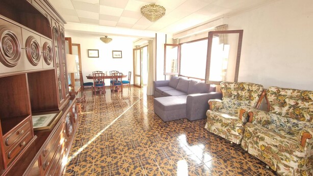 A spacious living area with a patterned floor, a sofa, a floral armchair, a dining table with chairs, and a cabinet. - 114m² 3 Bedroom Apartment with Balcony Lift and AC (Nules)