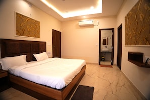 A bedroom with a bed, a nightstand, a wall-mounted air conditioner, a mirror, and a door leading to a bathroom. - Hotel Maruti Nandan (Varanasi)