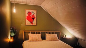 Ein Schlafzimmer mit schrägen Wänden, einem Bett mit zwei Kissen, einem gerahmten Kunstwerk an der Wand und einem Nachttisch mit Pflanze und Kerze.