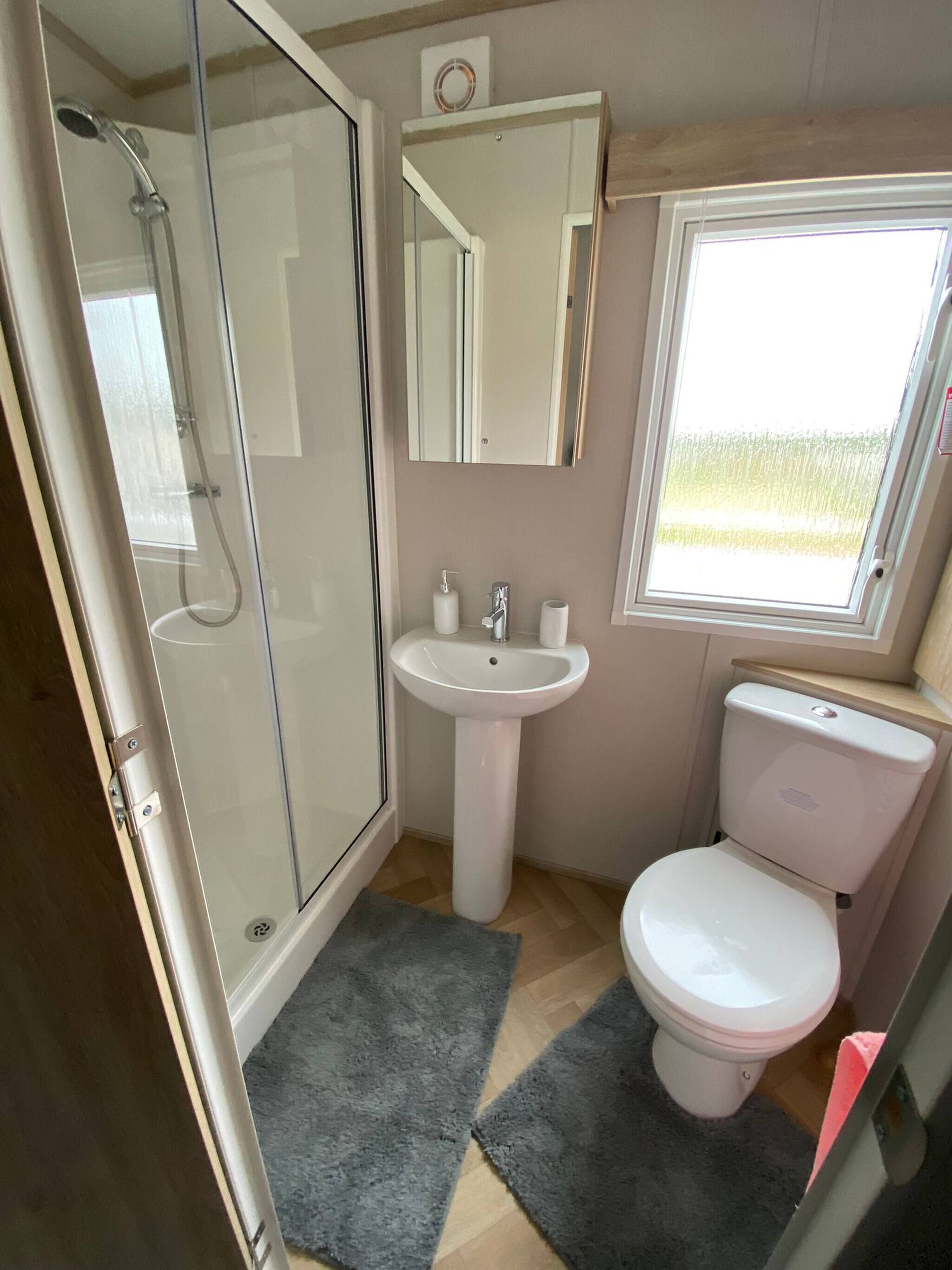 Un baño pequeño con mampara de vidrio, lavamanos pedestal, inodoro y ventana que proporciona luz natural.