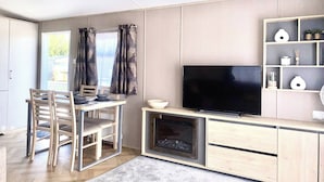 A modern living room with a TV, wooden furniture, a fireplace, and a window with curtains. - Elegant 2 Bed One and Half Bath Caravan in Holiday Resort - Sleeps 6 (Great Billing)