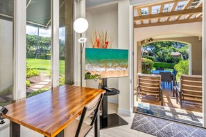 A room with a wooden table, a painting of a beach scene, and a view of a garden through a glass door. - The Palms at Wailea 1805 - Luxury 2BR condo, large lanai, Full AC, Kitchen, WiFi (Kihei)