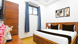 A hotel room with a wooden bed, a flat-screen TV, an air conditioner, a window with blue curtains, and three framed wall art pieces. - Hotel O Mana Camp Near Airport (Raipur)