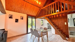 A spacious room with a wooden ceiling, a glass table with chairs, a staircase, and a view through a glass door. - The Campagnarde (Flers)