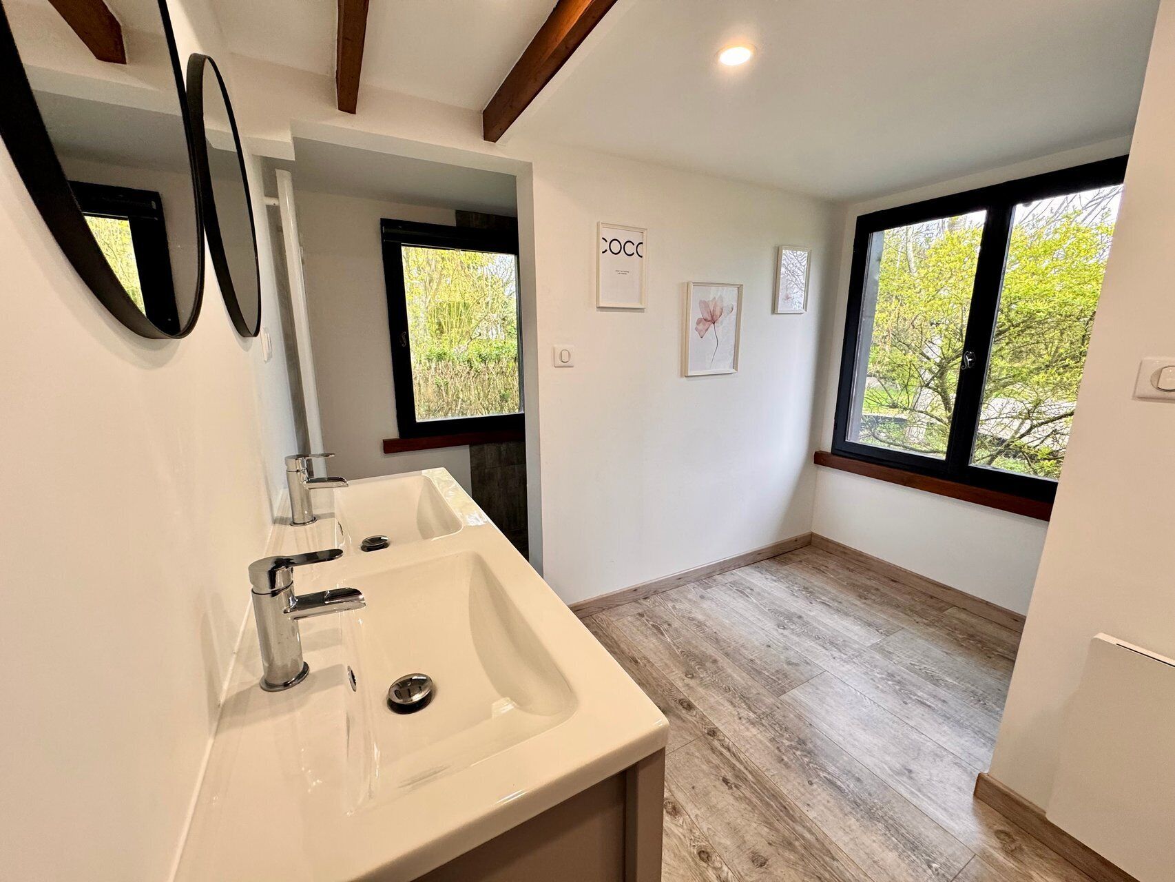 Une salle de bain moderne avec un grand lavabo, deux miroirs, une fenêtre et une porte donnant sur une autre pièce.