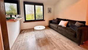 A living room with a dark brown sofa, a white coffee table, a window with a view of trees, and a framed picture on the wall. - The Campagnarde (Flers)