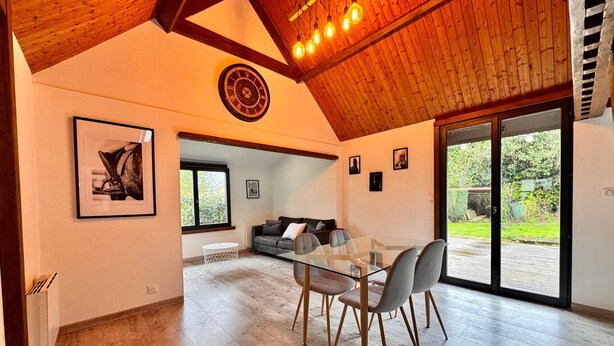 A spacious living area with a glass dining table, grey chairs, a wooden ceiling, and a large window offering a view of the outdoors. - The Campagnarde (Flers)