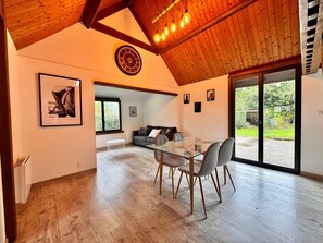 A spacious living area with a glass dining table, grey chairs, a wooden ceiling, and a large window offering a view of the outdoors. - The Campagnarde (Flers)