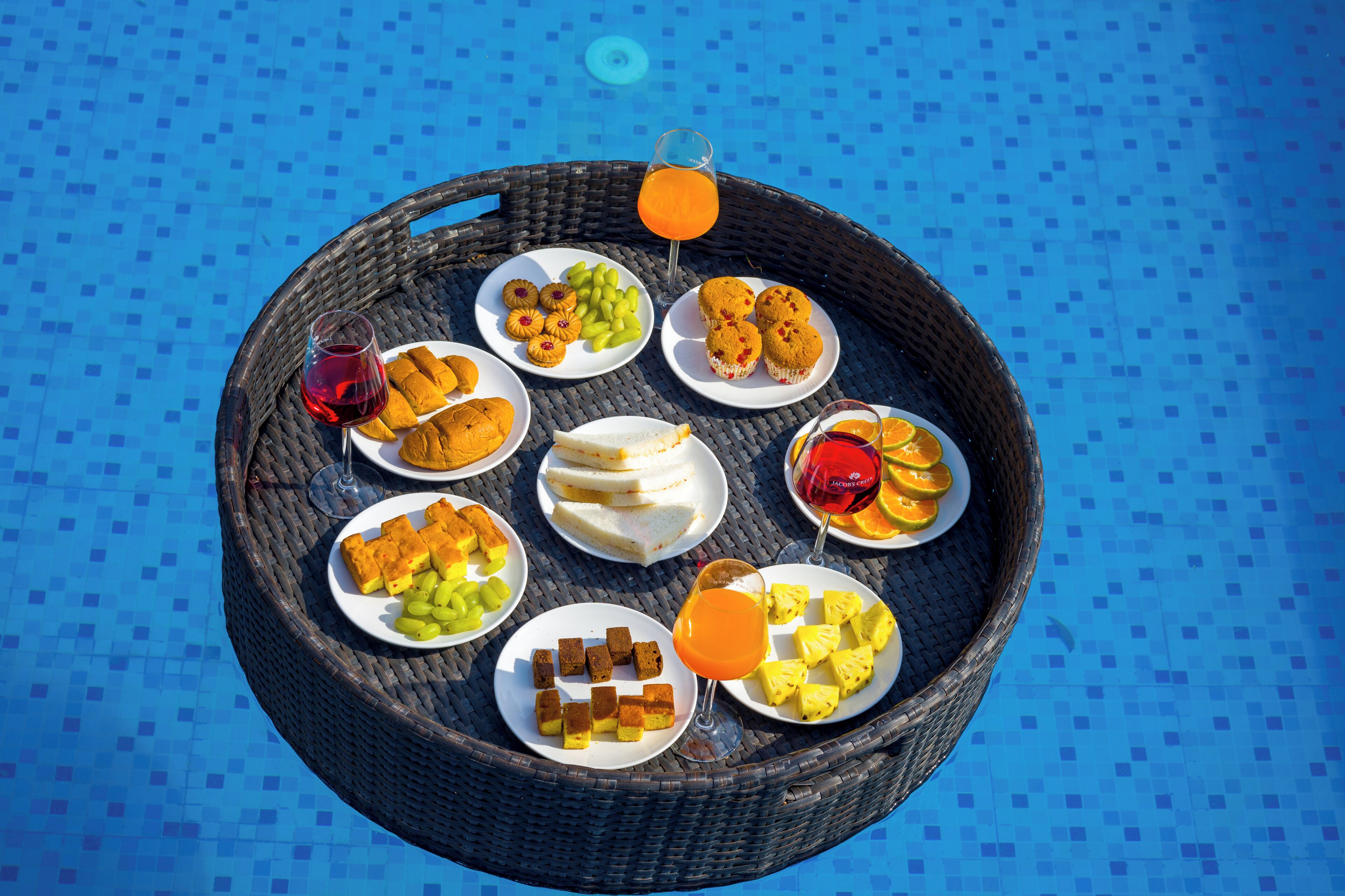 A round wicker tray with various food items and drinks on a blue tiled surface.