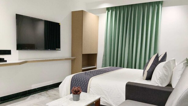 A hotel room with a bed, a sofa, a TV mounted on the wall, a small table, and a window with curtains. - Hotel Niyathi (Erode)