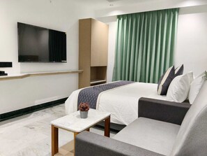 Quarto de hotel com cama, sofá, televisão na parede, mesa pequena e janela com cortinas.