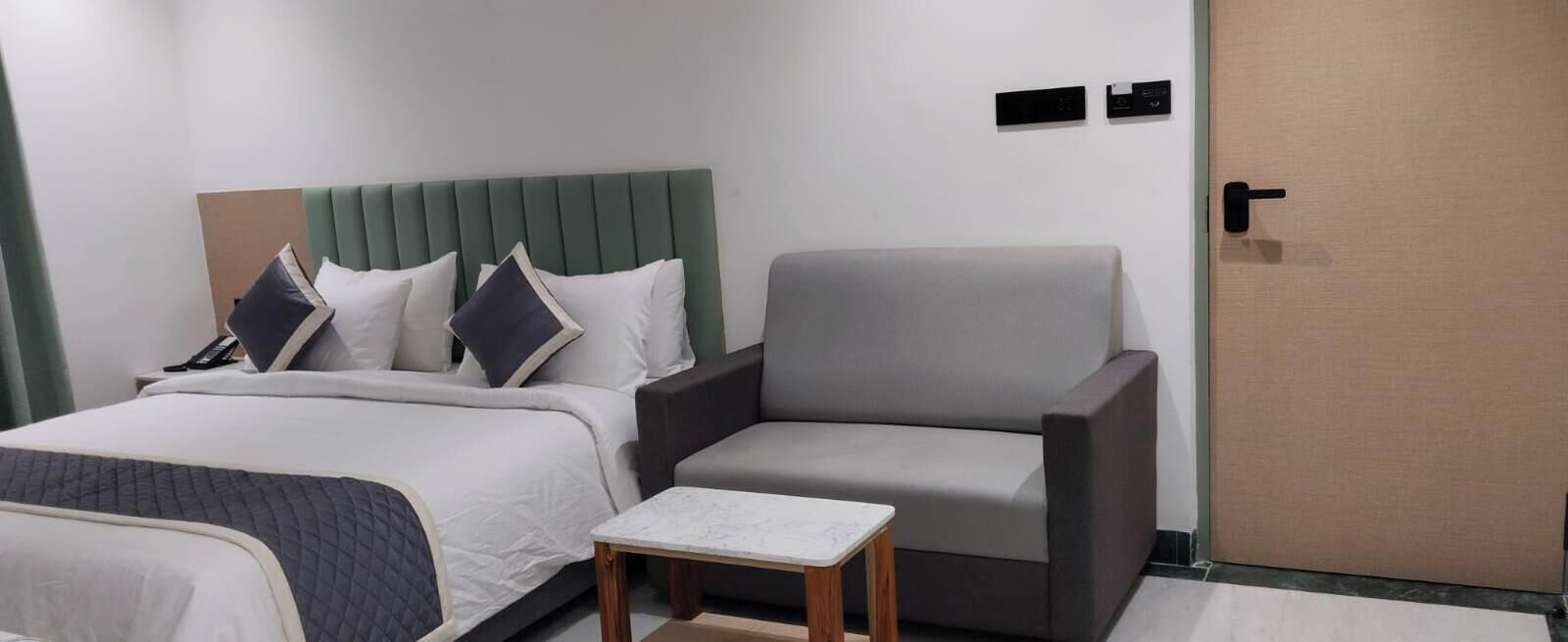Quarto de hotel com uma cama, uma poltrona cinzenta, uma mesinha e uma porta.