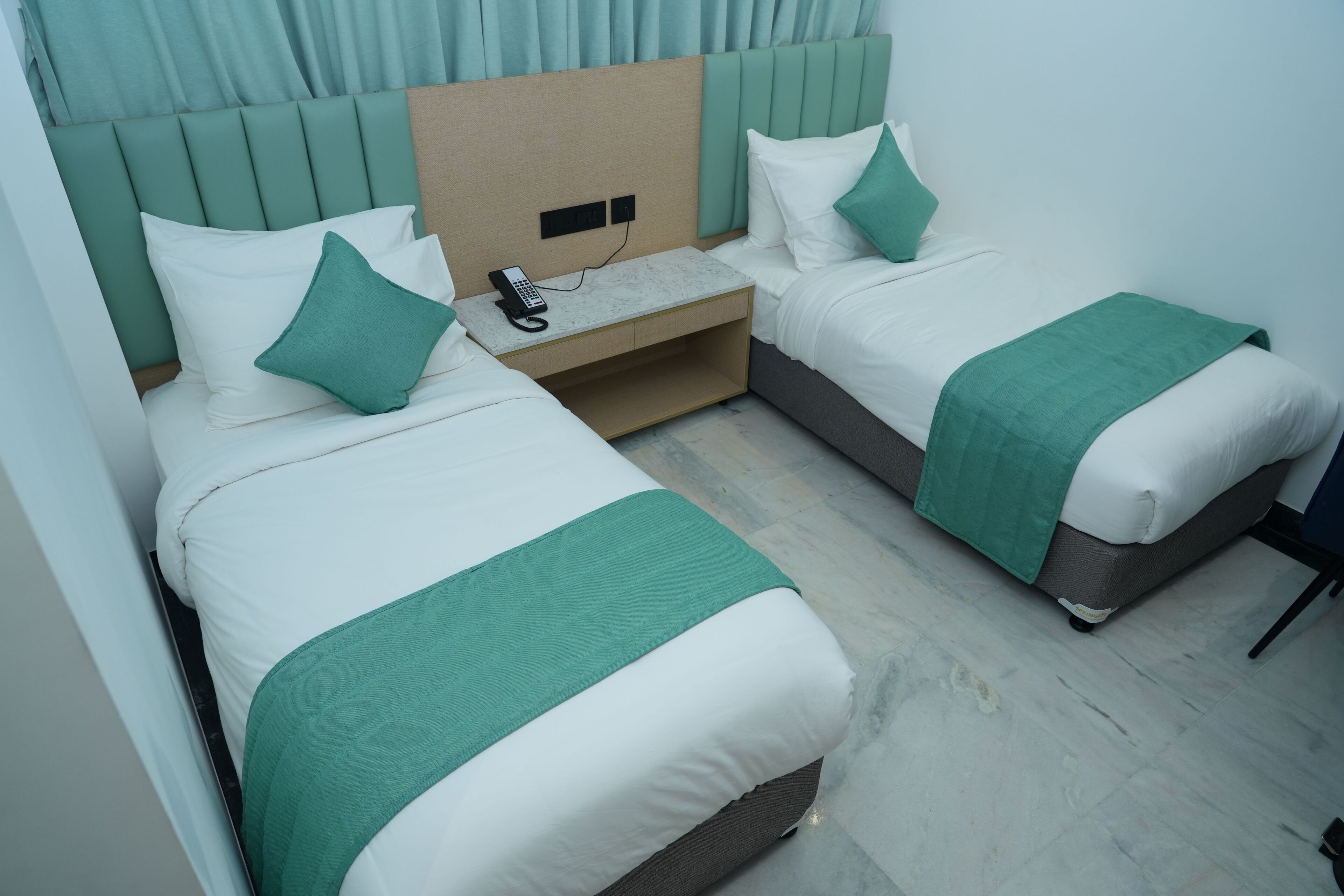 Habitación de hotel con dos camas individuales, una mesita de noche pequeña con teléfono y cabecero con estante incorporado.