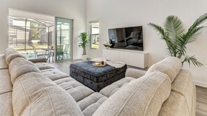 Ein modernes Wohnzimmer mit einem großen Ecksofa, einem Couchtisch, einem flachbildfernseher an der Wand und einer Schiebeglastür, die auf eine Terrasses führt.