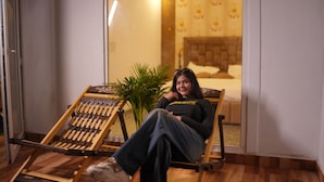 A person sitting on a wooden lounge chair in a room with a bed, a potted plant, and a window. - Sundown Resort (Noida)