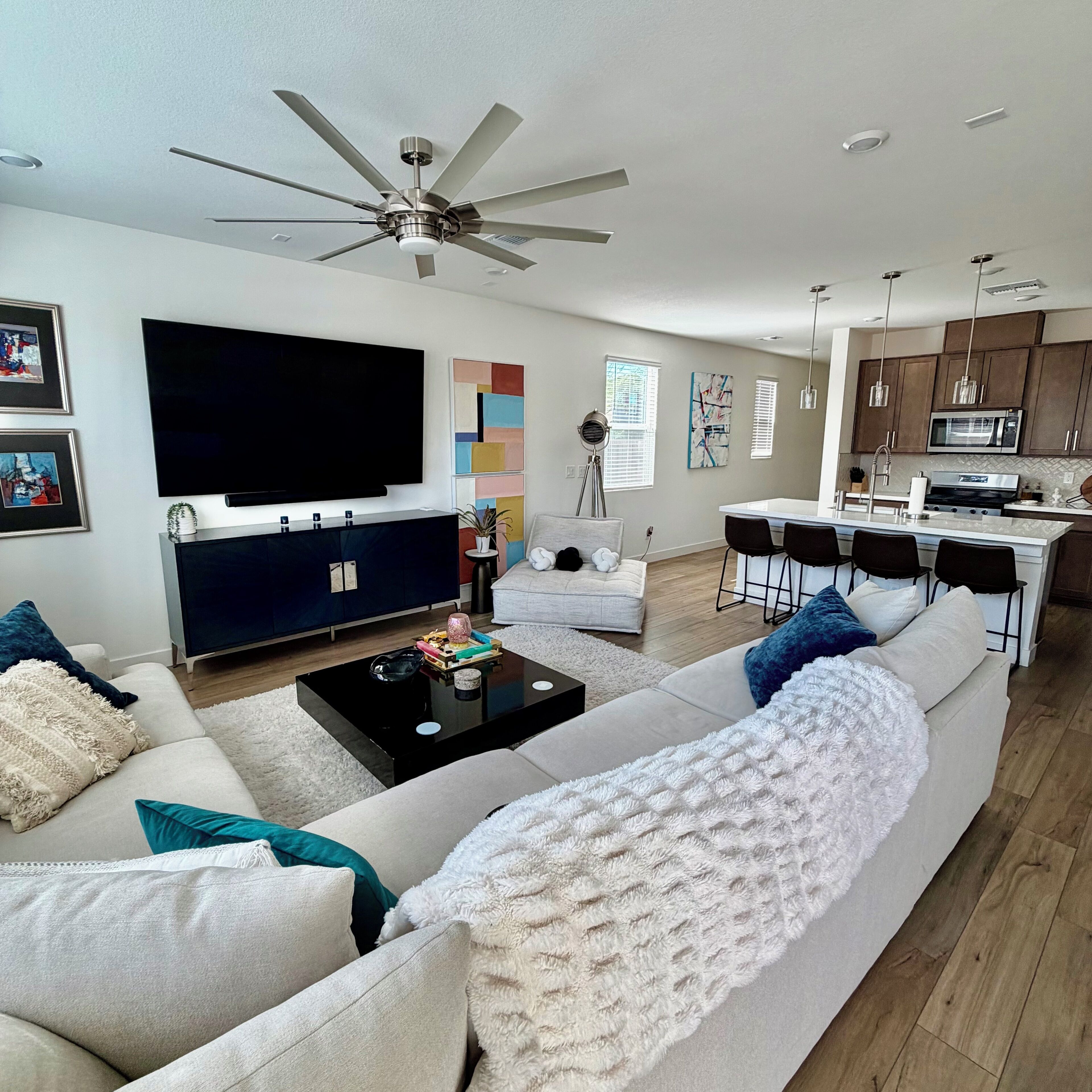 Una sala moderna con un televisor de pantalla plana grande, un ventilador de techo, una zona de comedor con taburetes altos y una cocina con microondas.