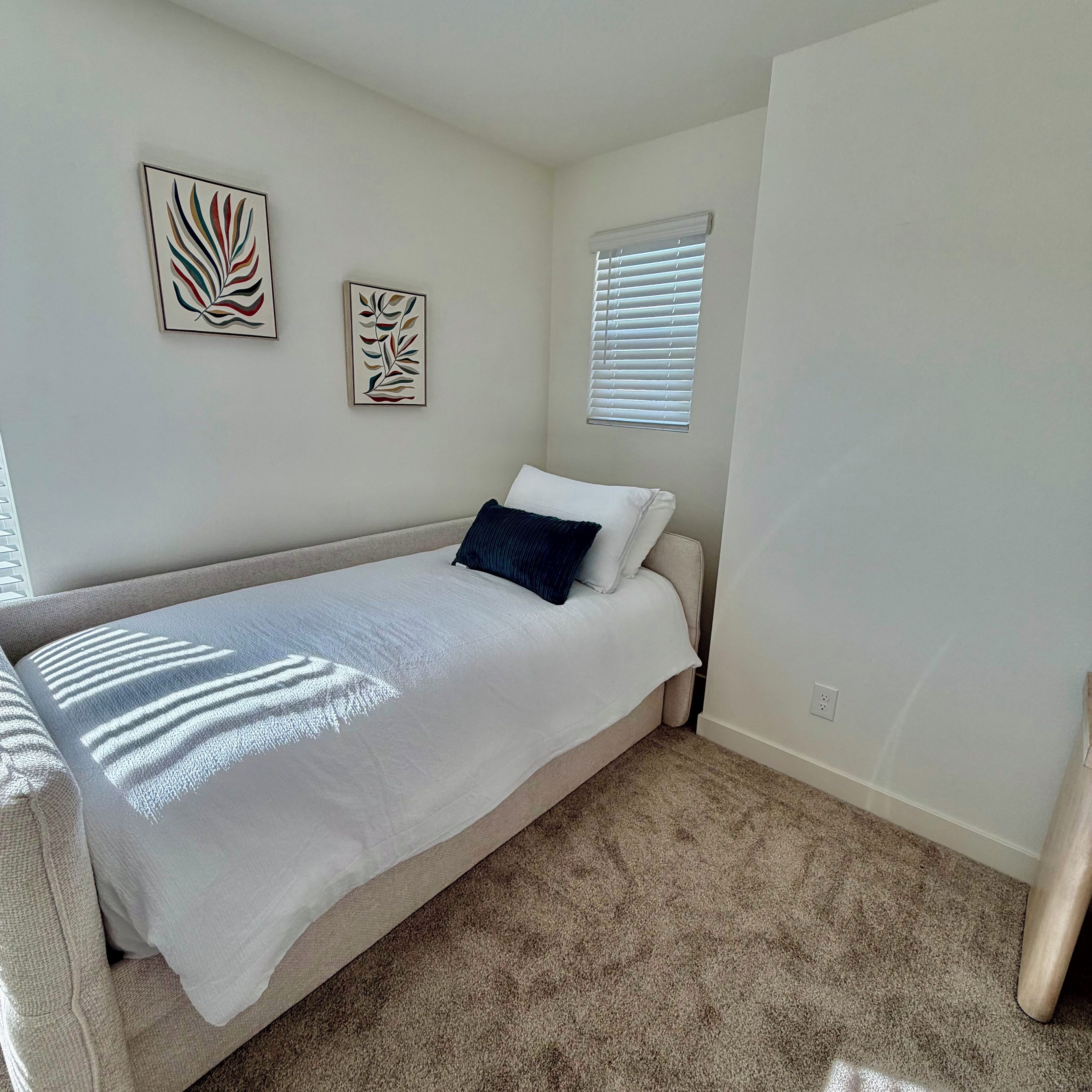 A bedroom with a bed, two wall art pieces, a window with blinds, and a carpeted floor.