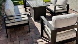 Eine Terrasse im Freien mit Tisch und vier Stühlen, einem Backsteinboden und einem Rasenstück im Hintergrund mit Mauer und Fenster.