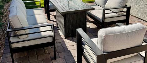 Een terras buiten met een tafel en vier stoelen, een stenen vloer en een gazon met een muur en raam op de achtergrond.