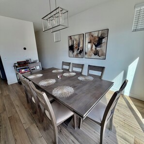 Een eetruimte met een rechthoekige tafel, zes stoelen en decoratieve kantdoekjes. Aan de muur hangen abstracte schilderijen en erboven bevindt zich een hanglamp.
