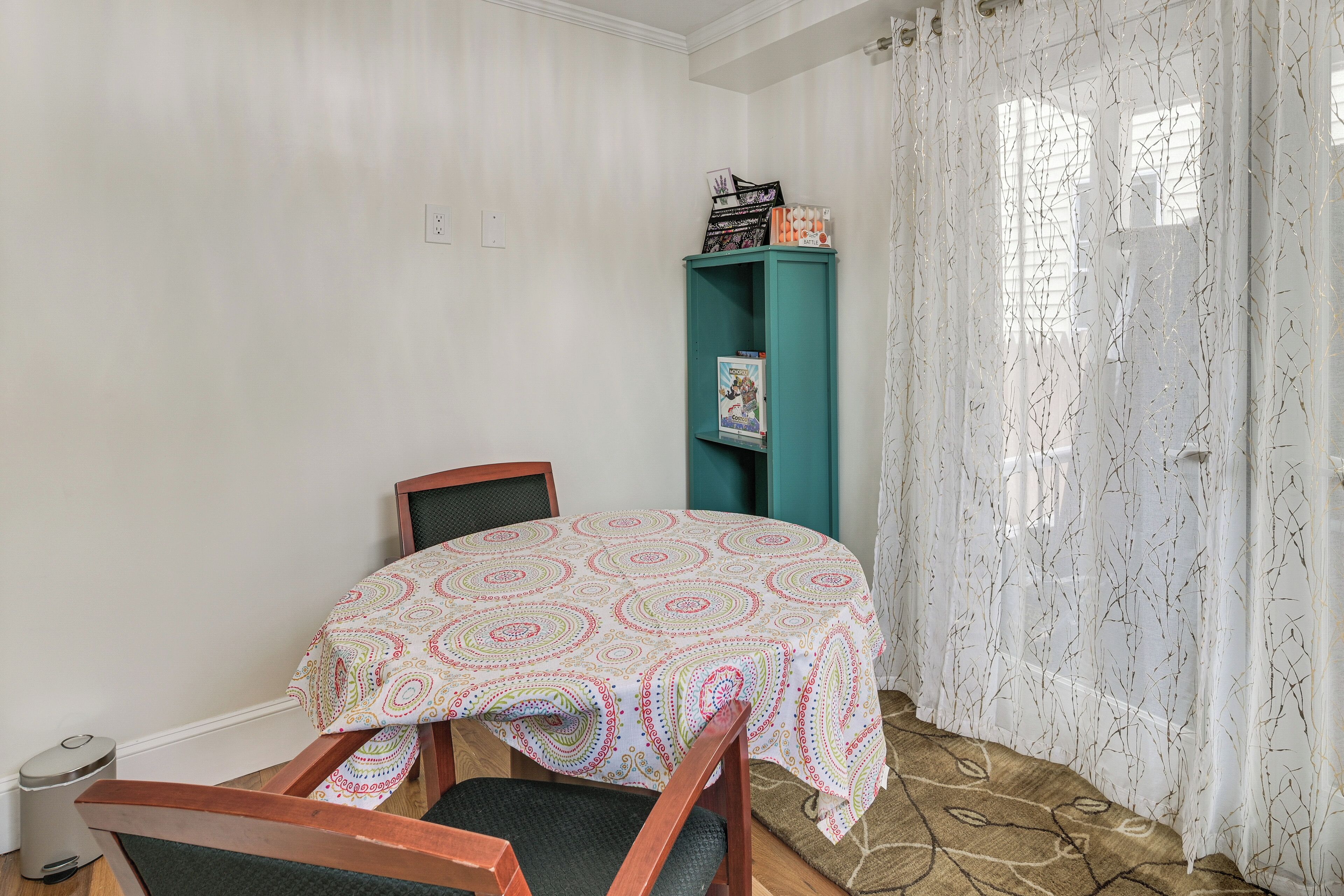 A small room with a bed, a chair, a teal bookshelf, and a window with sheer curtains.