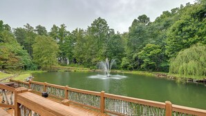 A wooden deck with a railing overlooking a pond with a fountain, surrounded by lush greenery. - 2BD Mtn Retreat near Nantahala w/ Firepit and Pond (Andrews)