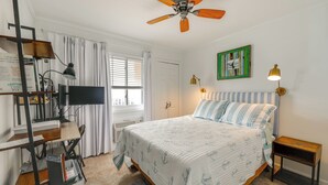 Un dormitorio con cama, ventilador de techo, televisión, escritorio con computadora y estantería.