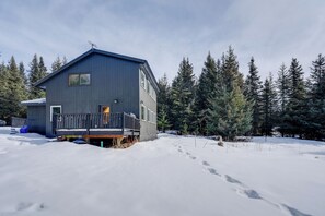 Una cabaña de dos pisos con balcón, rodeada de nieve y árboles de hoja perenne.