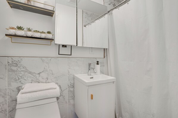 A bathroom with a white sink, marble tiles, a towel rack, and a shower curtain. - Easy Access to NYC! Sleek Suburban Apartment (Mamaroneck)