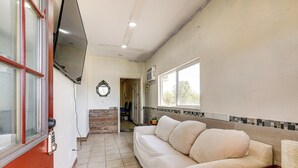 A room with a beige sofa, a TV mounted on the wall, a door with a red frame, and a window with a view of the outside.