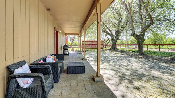 An outdoor covered patio with wicker furniture, a grill, and a view of trees and a fence.
