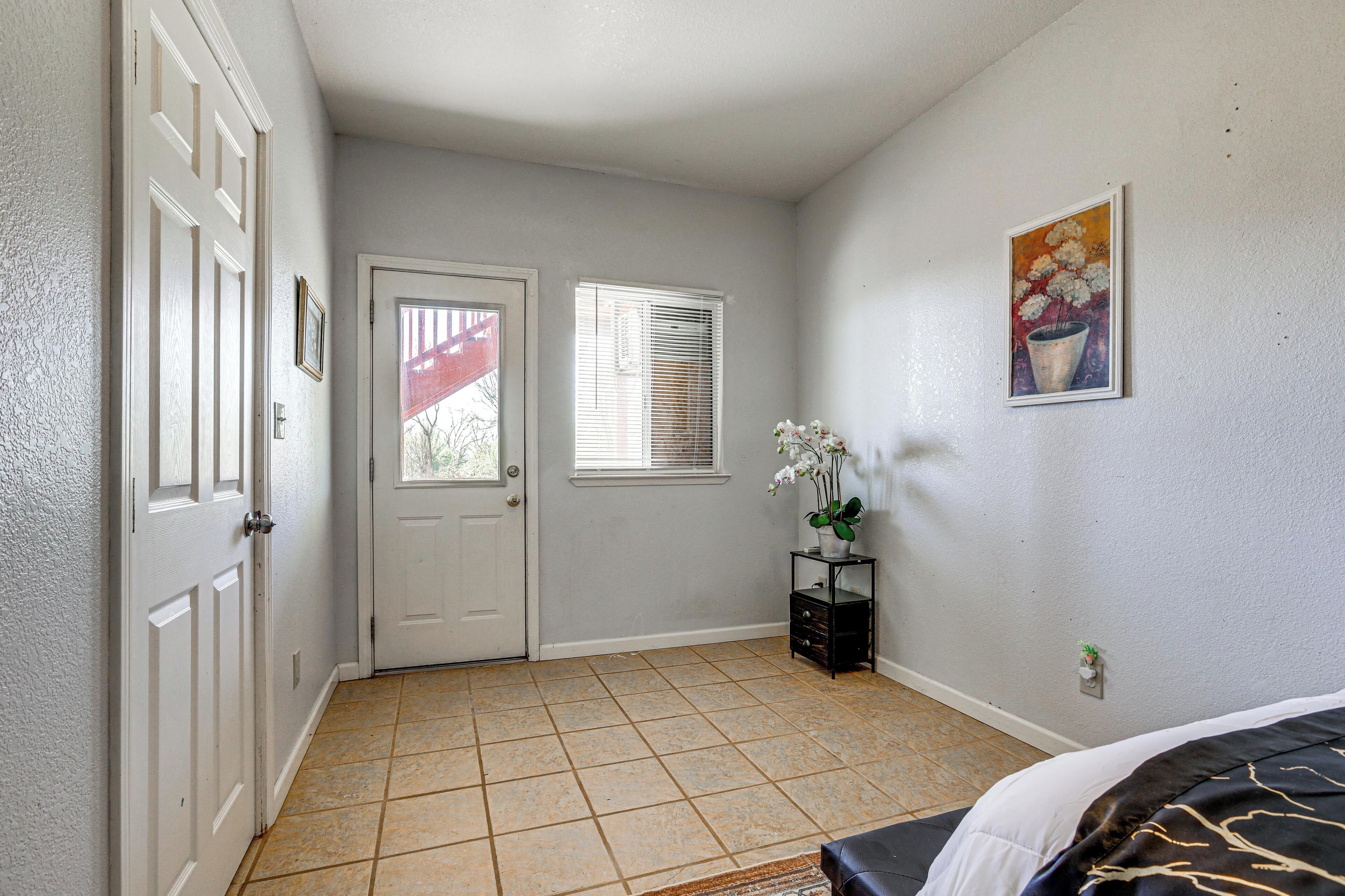 A room with a door, a window with blinds, a small plant on a stand, and a painting on the wall.