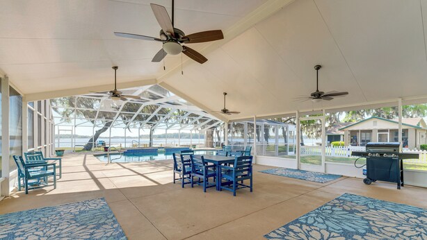 A covered outdoor area with a pool, blue chairs, a table, ceiling fans, and a grill. - Lake Days + Pool Fun! Nature Coast Retreat (Hernando)