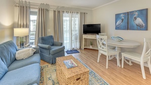 Ein Wohnzimmer mit einer Couch, einem Sessel, einem Couchtisch, einem Fernseher, einem Esstisch mit Stühlen und Kunstwerken an der Wand.
