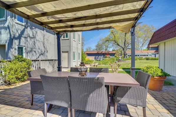 Un patio al aire libre con una mesa y sillas, una pérgola en la parte superior y vistas de un edificio y jardín.