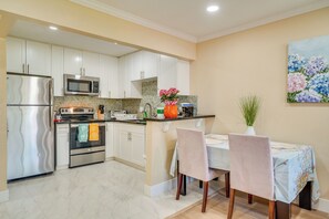 Una cocina con armarios blancos, electrodomésticos de acero inoxidable, un comedor con mesa y sillas, y un cuadro en la pared.