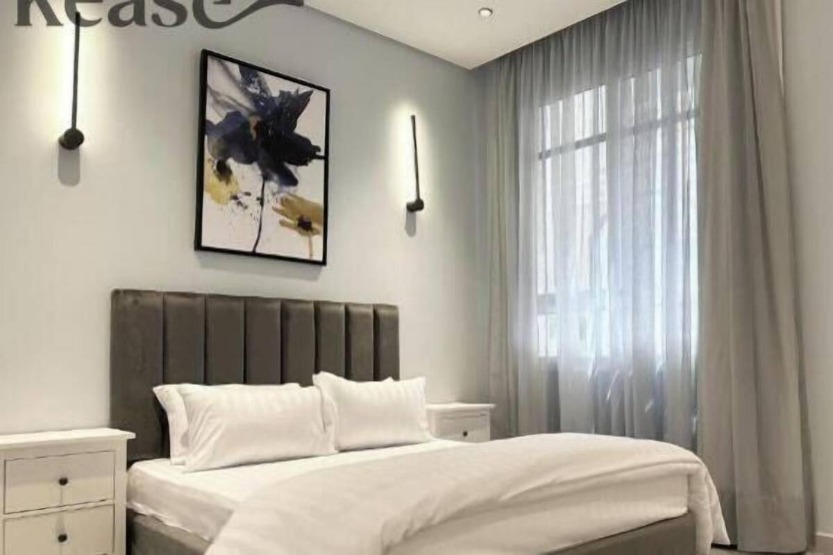 A bedroom with a bed, a nightstand, a wall-mounted light, a window with curtains, and a painting.