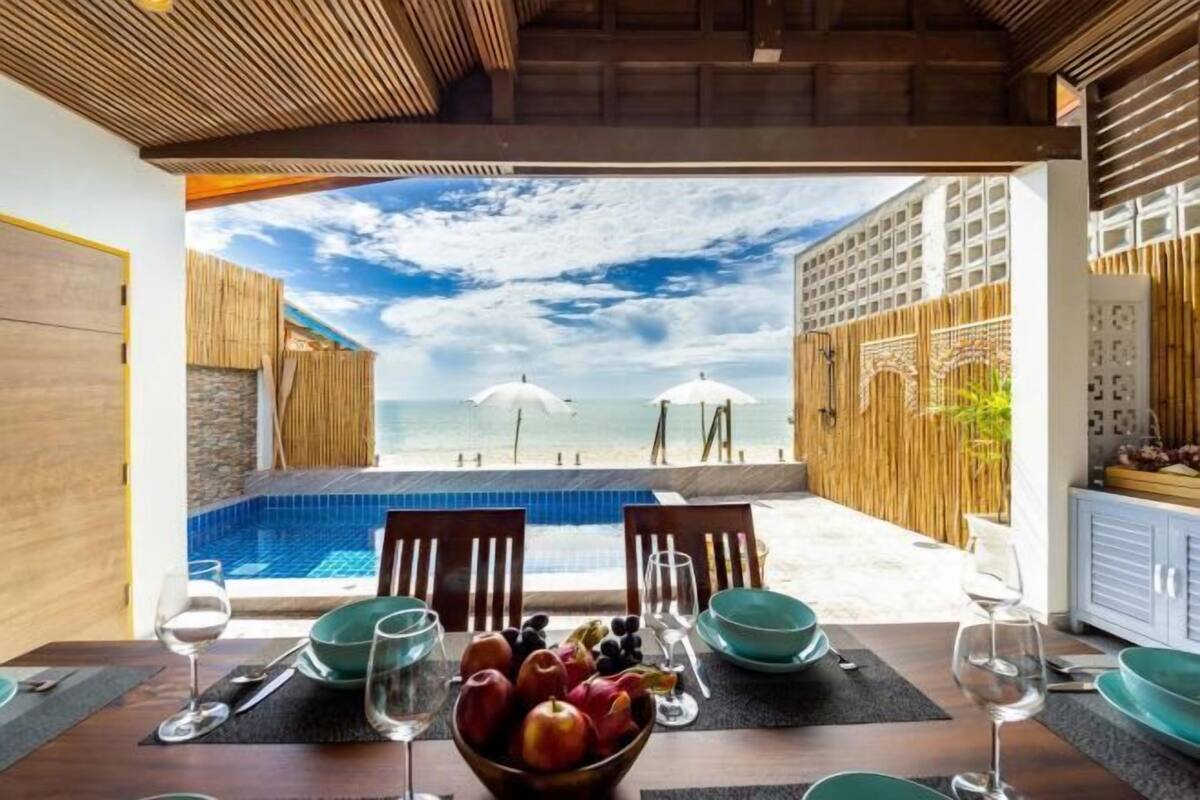 A dining area with a view of a pool and beach umbrellas, wooden walls, and a table set with plates and glasses.