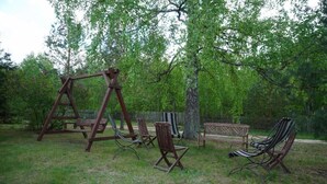 A wooden swing set, a bench, and several chairs in a grassy outdoor area with trees in the background. - Olszówka (Goniądz)