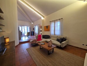 Ein großzügiger Wohnraum mit einer Couch, einem Couchtisch und einem Fernseher. Eine Tür führt in einen weiteren Raum, ein Fenster mit Vorhängen ist ebenfalls vorhanden.