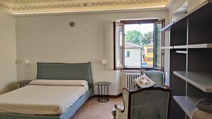 Ein Schlafzimmer mit Bett, Sessel, kleinem Tisch, Heizkörper und Fenster mit Blick auf Gebäude.