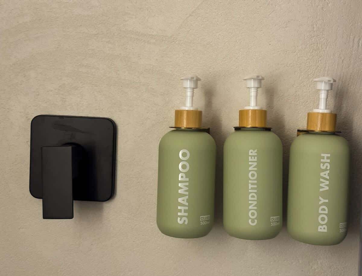 Dispensador montado en la pared con tres botellas etiquetadas 'SHAMPOO', 'CONDITIONER' y 'BODY WASH'.