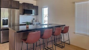A modern kitchen with a central island, bar stools, stainless steel appliances, and a window with blinds.