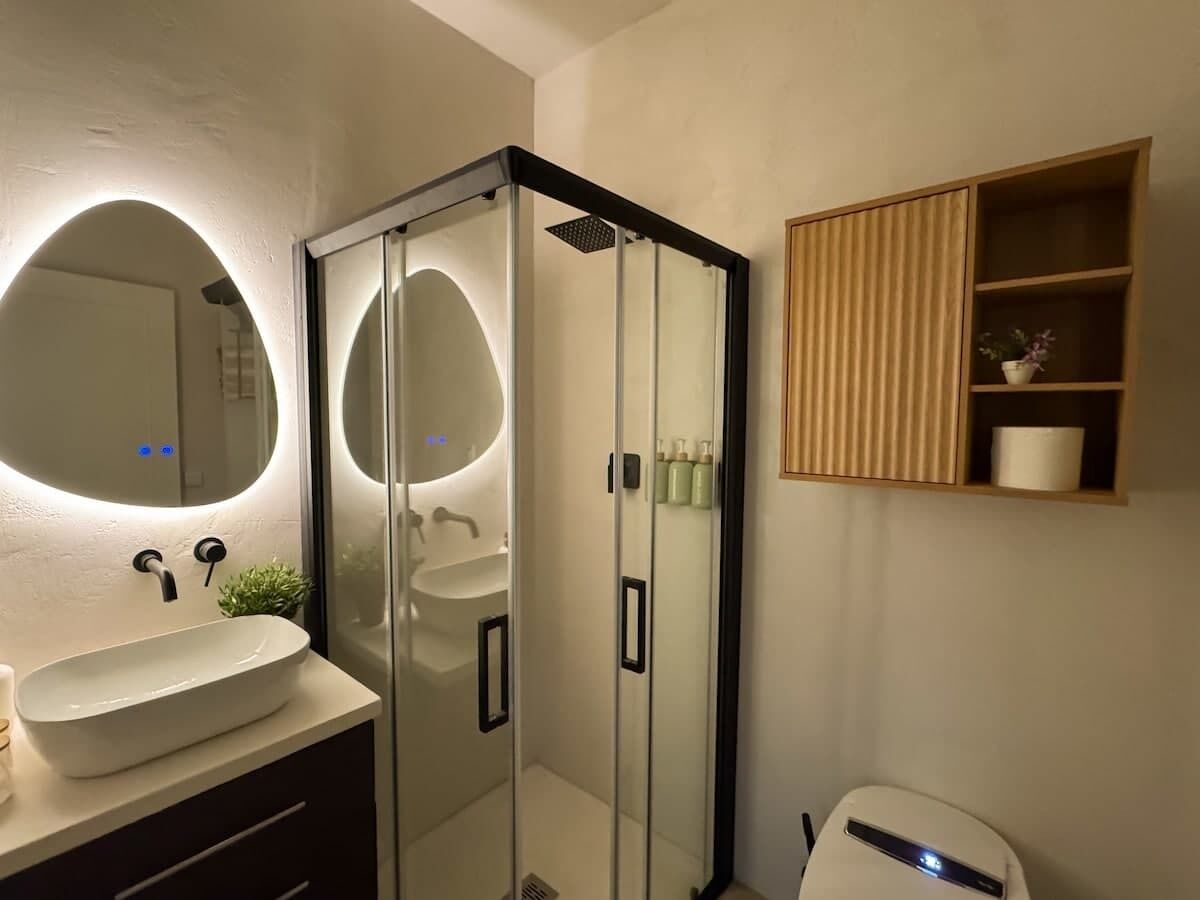 A bathroom with a glass-enclosed shower, a white sink, and a mirror with a circular light.