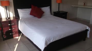 Una cama bien hecha con edredón blanco y almohada roja, flanqueada por dos mesitas de noche con lámparas, en una habitación con suelo de baldosas.