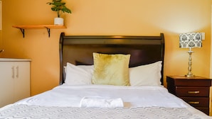 Een net opgemaakt bed met een bedsprei met een patroon, een nachtkastje met een lamp, en een plankje met een plant.