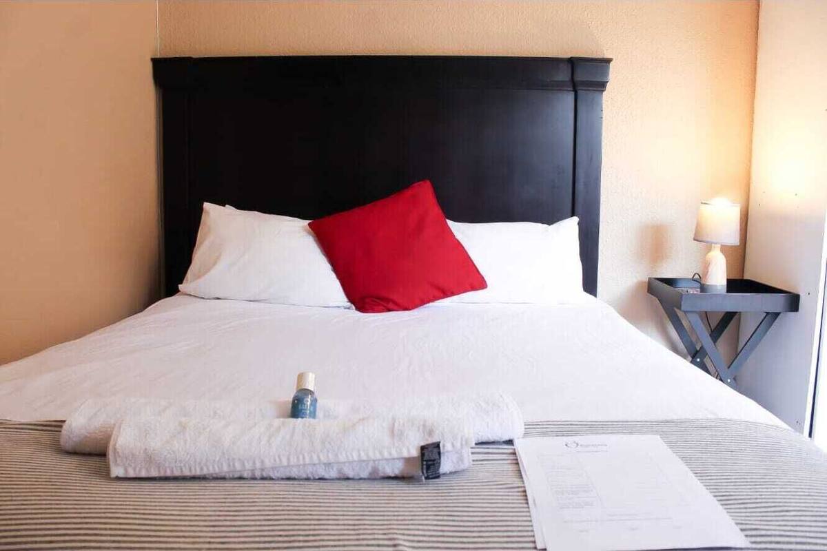 Ein ordentlich gemachtes Bett mit weißen und roten Kissen, einer weißen Decke und einer kleinen Flasche darauf. Ein Lampenschirm steht auf einem Nachttisch neben dem Bett.