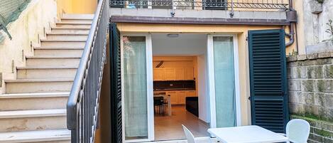 Un edificio residencial con una escalera que conduce a un balcón, una puerta corrediza de vidrio que da a una cocina y una pequeña zona de estar al aire libre con una mesa y sillas.