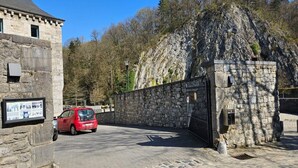 A stone building with a car parked in front, a gate, and a rocky hillside in the background.
